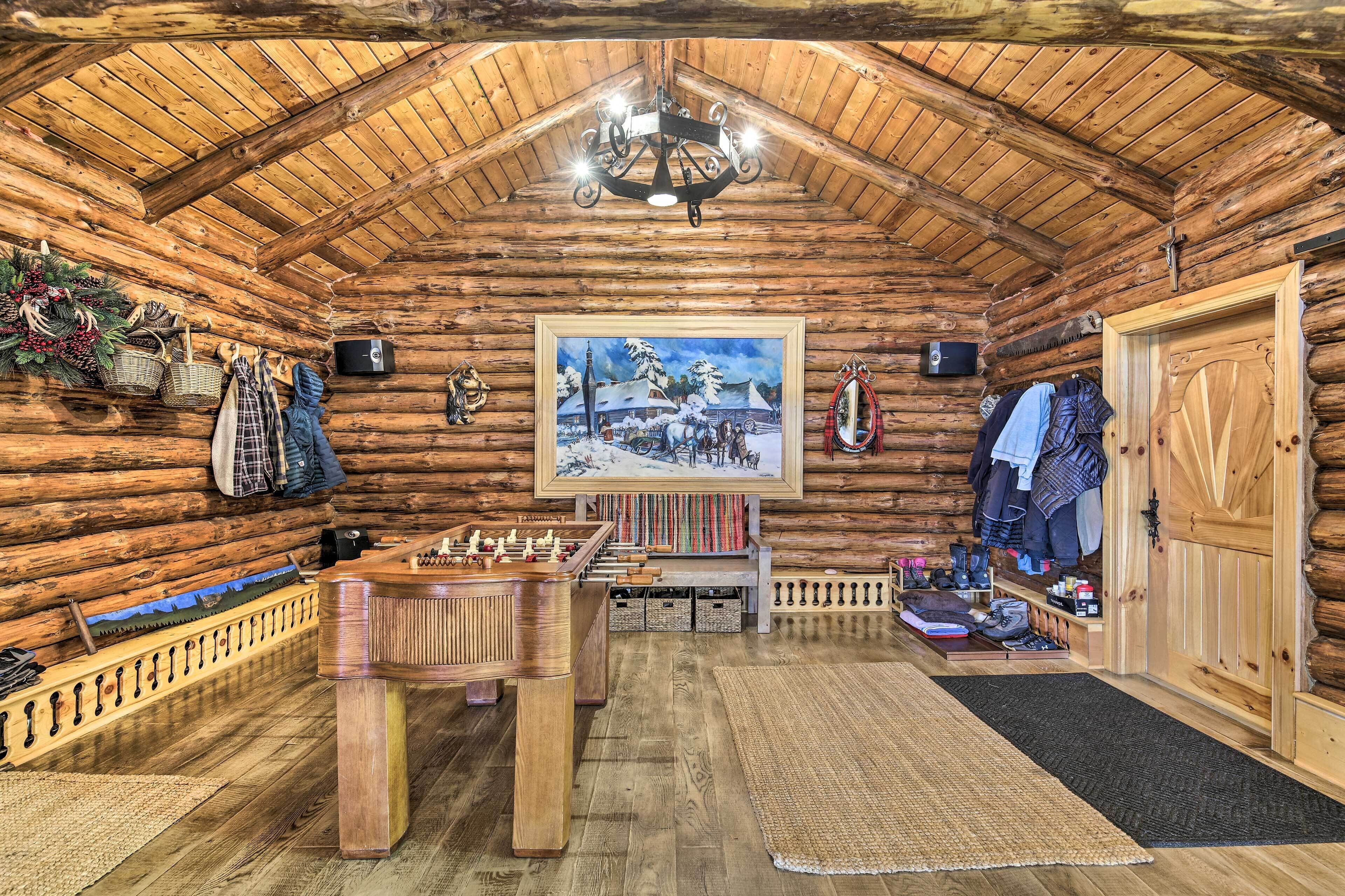 A log cabin interior with a wooden ceiling, a painting of a snowy landscape, a foosball table, and a clothing rack.