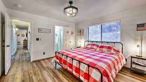 Une chambre avec une couvre-lit à carreaux, un ventilateur de plafond, une fenêtre avec des stores et une porte donnant sur une autre pièce.