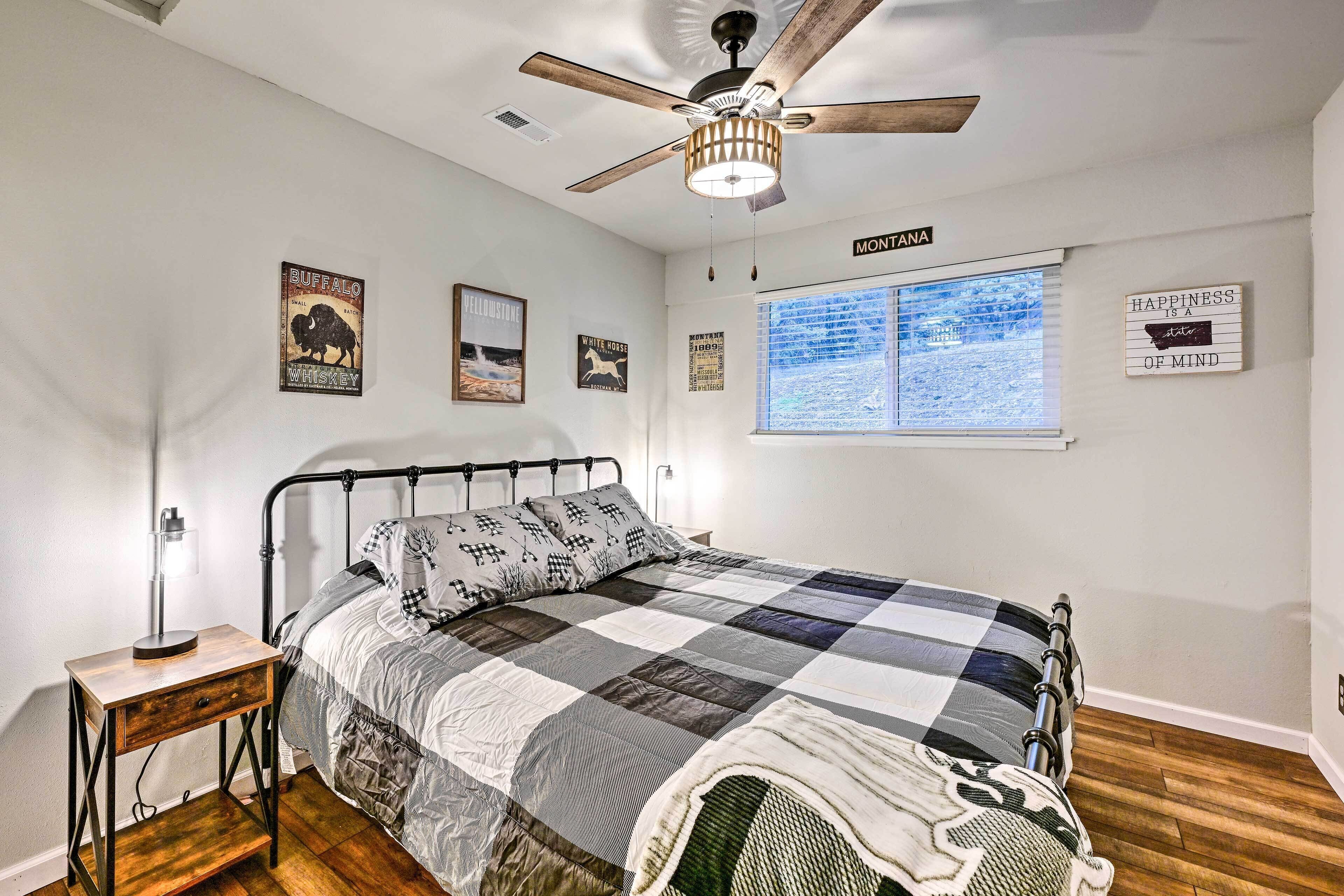 A bedroom with a metal bed frame, a quilted bedspread, a ceiling fan, a window with blinds, and wall art including a sign that says "HAPPINESS IS A STATE OF MIND".