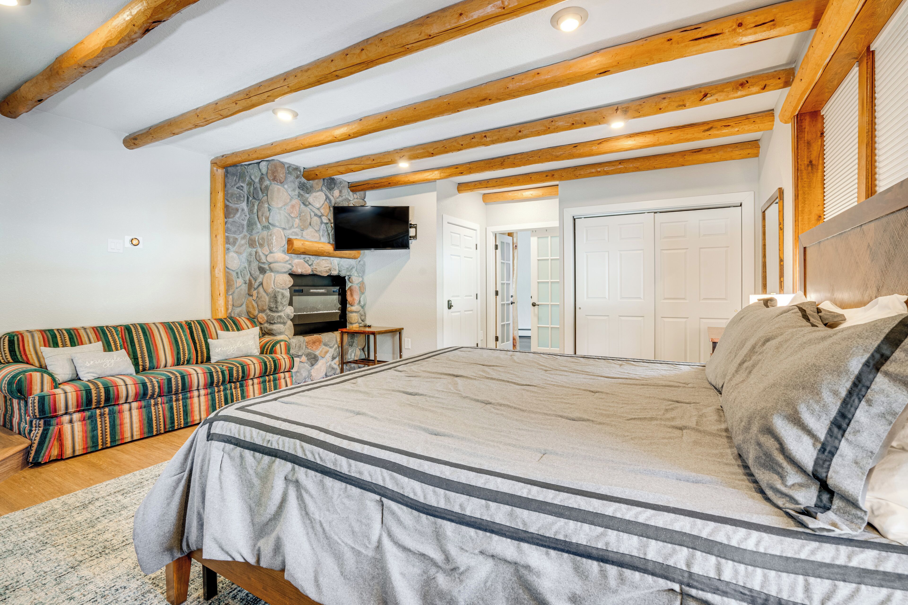 A spacious bedroom with a large bed, a stone fireplace, a colorful striped sofa, and wooden beams on the ceiling.