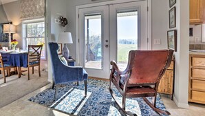 A dining area with a table set for four, a blue chair, a brown rocking chair, and a view of the outdoors through a door. - Family Farmhouse on 160-acre Ranch w/ Hiking Trail (Lawrenceburg)
