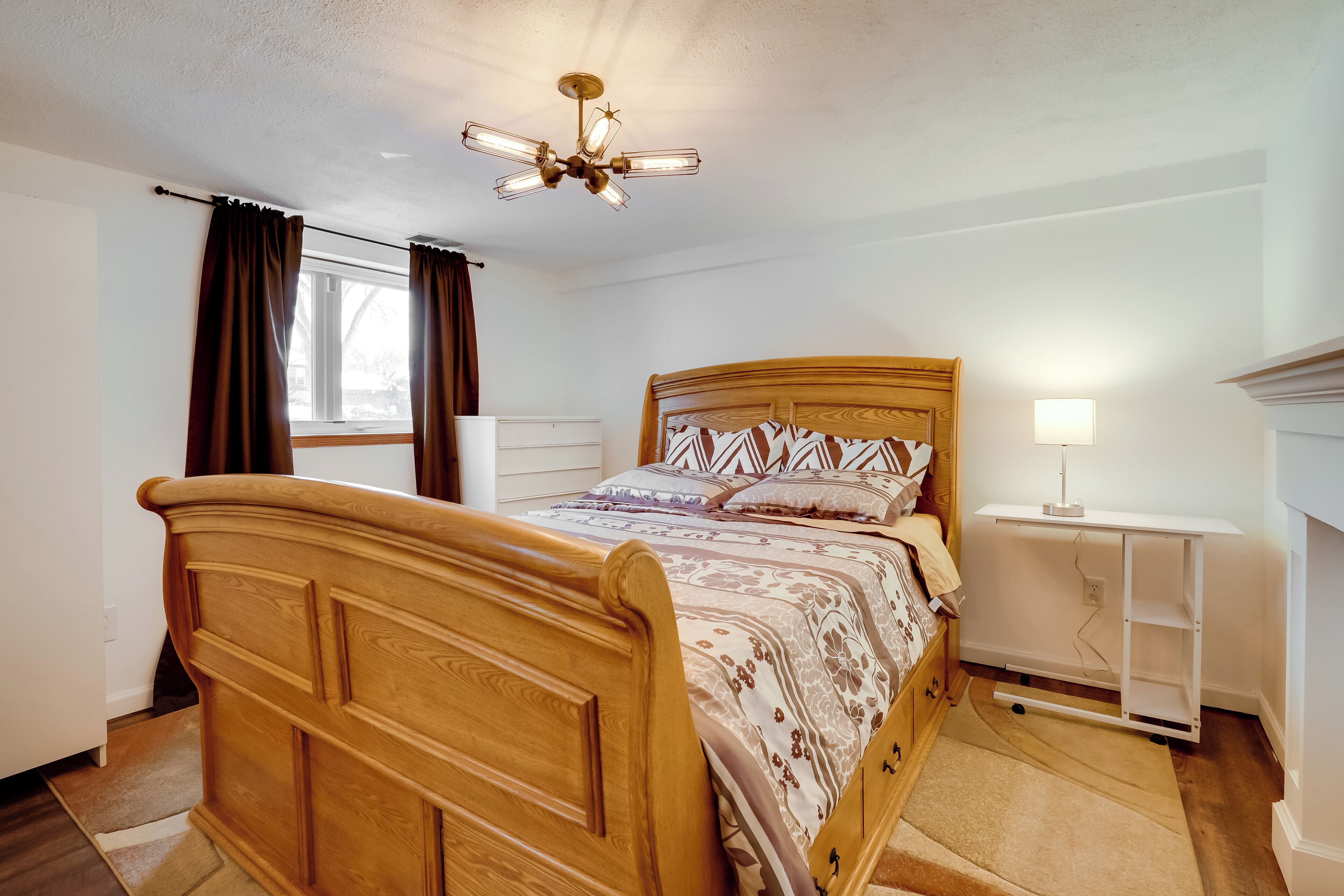 A bedroom with a wooden bed, a ceiling fan, a bedside table with a lamp, and a window with curtains.