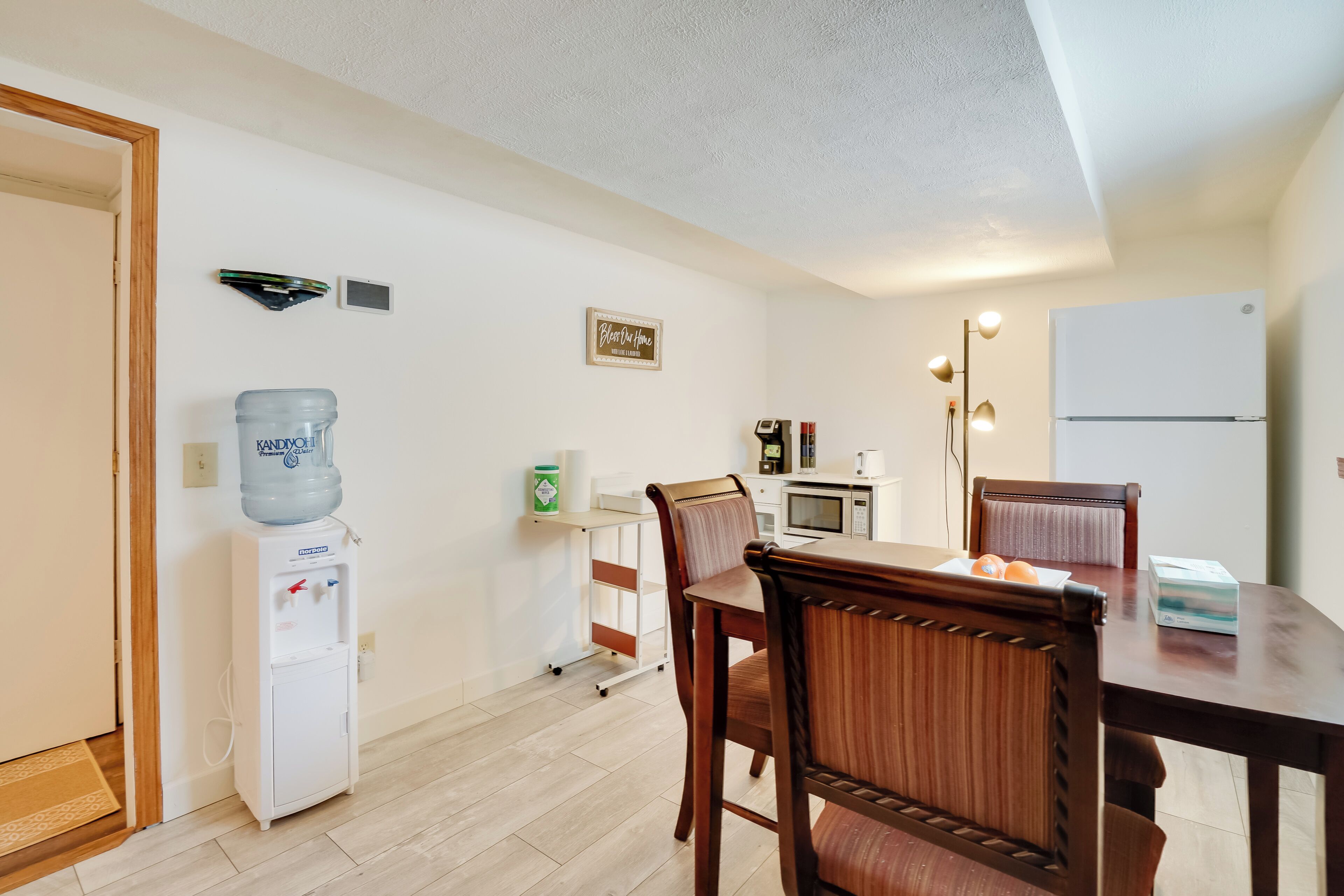 Een compacte keuken met een koelkast, magnetron en een waterdispenser. Er is een eettafel met stoelen en een klein kastje.