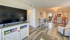 A spacious living room with a flat-screen TV, a dining area with a table and chairs, and a kitchen area with a sign reading "Emerald Isle". - Emerald Isle Vacation Rental w/ Resort Pool! (Emerald Isle)