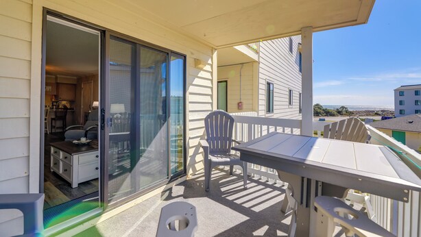 A covered outdoor area with a table and chairs, a sliding glass door leading to an interior space, and a view of the beach. - Emerald Isle Vacation Rental w/ Resort Pool! (Emerald Isle)