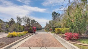 A paved pathway leading to a gated entrance with a brick wall and various shrubs and trees. - Emerald Isle Vacation Rental w/ Resort Pool! (Emerald Isle)