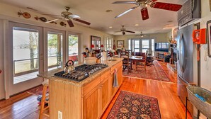 A spacious kitchen with a central island, wooden cabinets, and a large refrigerator. There are ceiling fans, a dining area with a rug, and a view through the glass doors. - Waterside Belhaven House & Cottage w/ Porch & Dock (Belhaven)