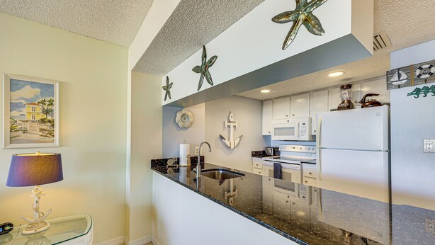 A kitchen with a granite countertop, white cabinets, and a refrigerator. There is a painting of a house on the wall, a lamp, and a decorative anchor. - Waterfront Condo w/ Pool Access in Punta Gorda! (Punta Gorda)