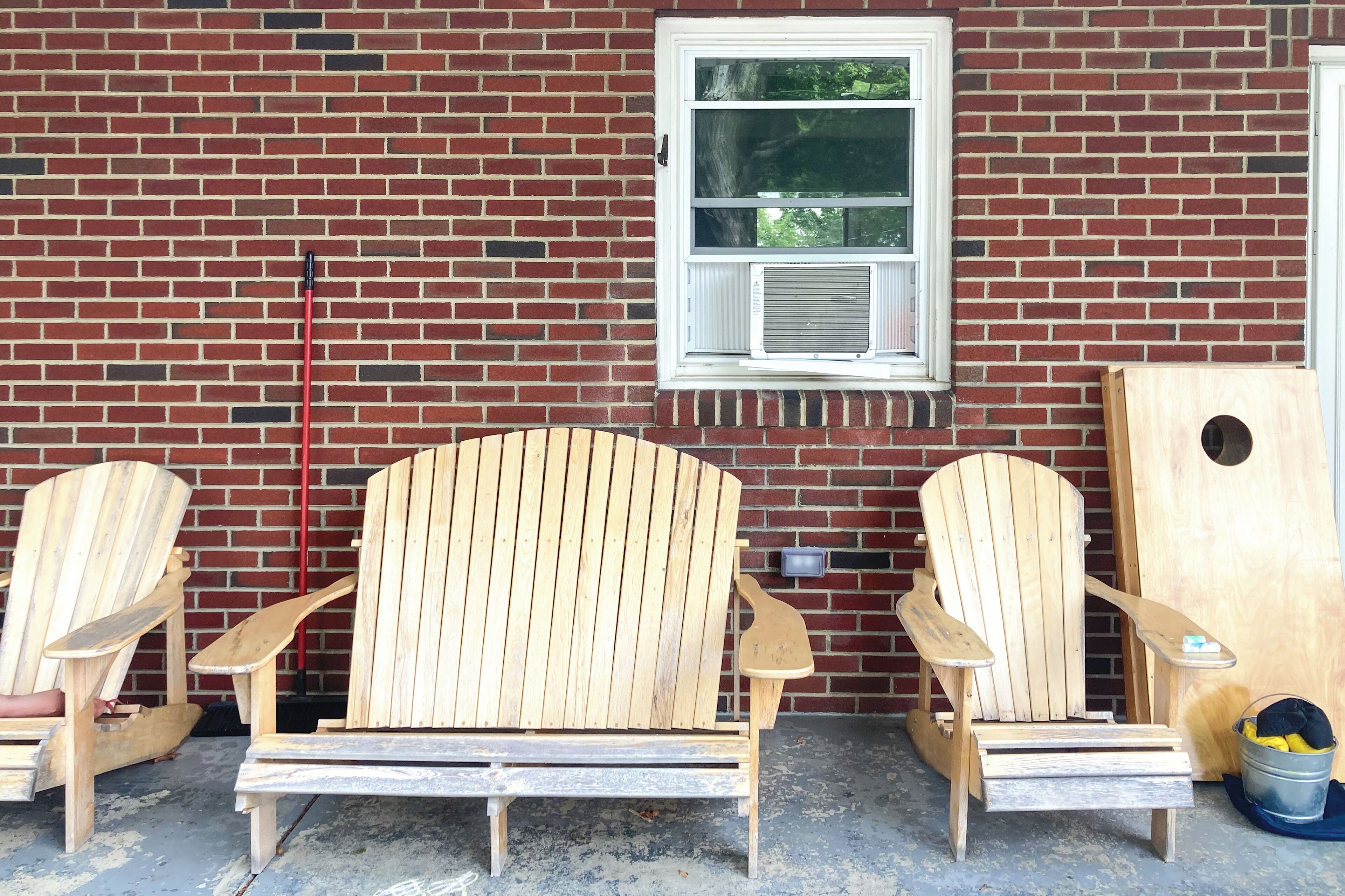 Tres sillas Adirondack de madera, un balde metálico y una puerta de madera frente a un muro de ladrillo.