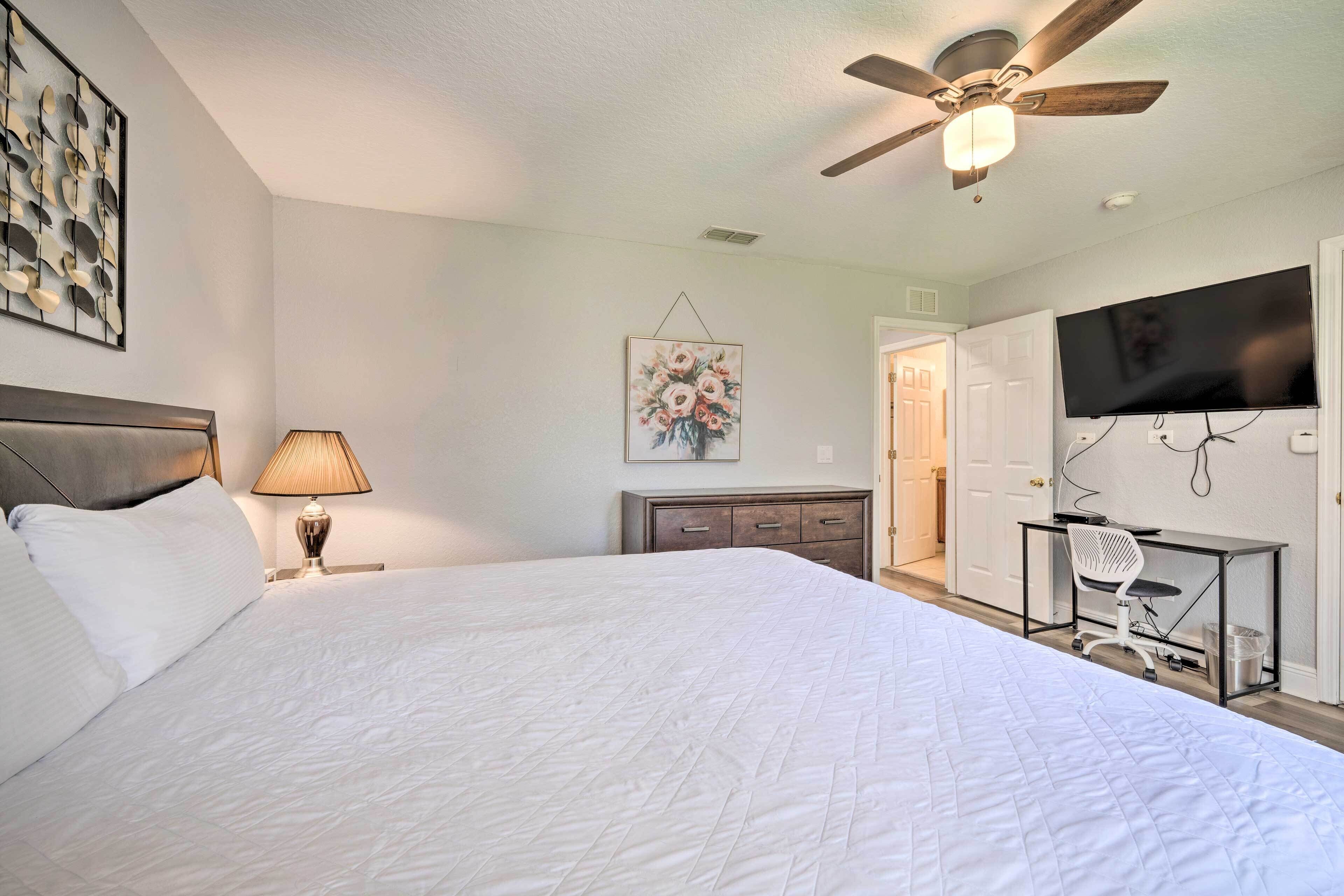 A bedroom with a large bed, a desk with a chair, a ceiling fan, a TV, and a door leading to another room.