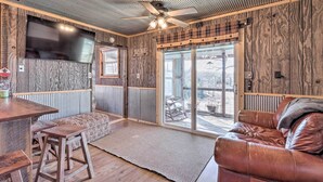 A cozy cabin interior with a wooden ceiling, a ceiling fan, a TV mounted on the wall, a large sliding glass door leading to a deck, and a leather couch. - Rustic Retreat - Steps to Pomme De Terre Lake (Flemington)