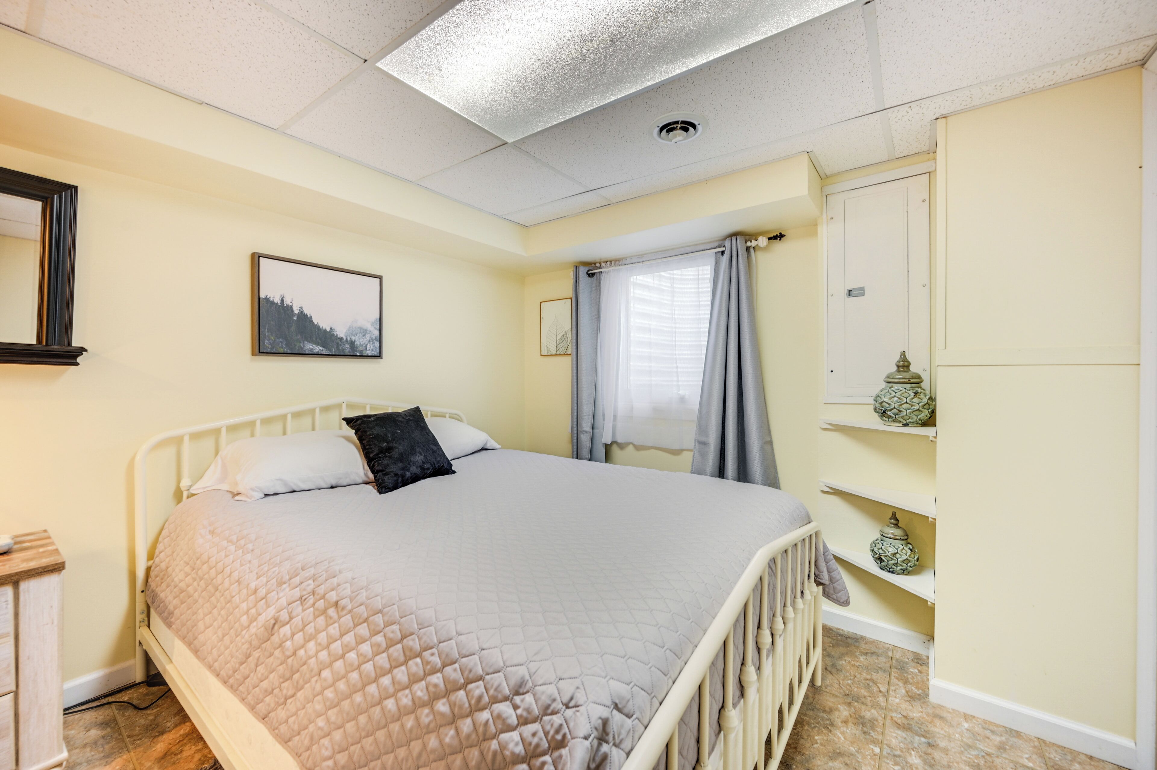 Un dormitorio con cama, mesita de noche, cómoda, ventana con cortinas y estante con objetos decorativos.