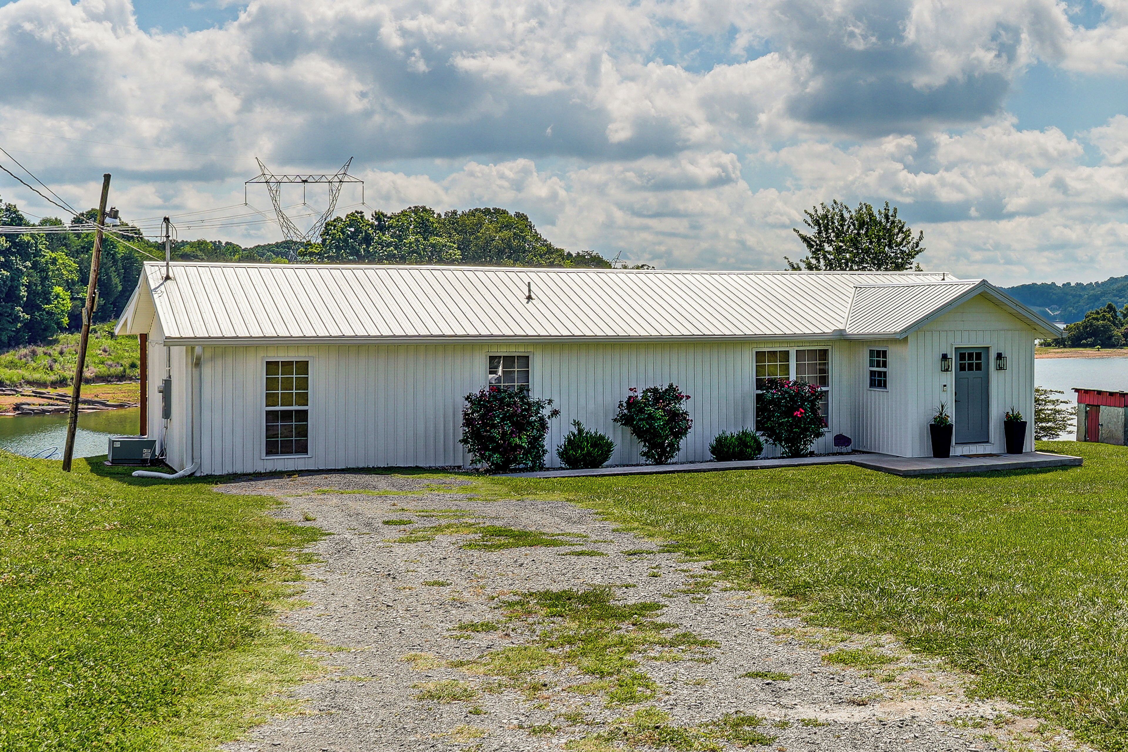 A white single-story building with a metal roof, a gravel driveway, and small bushes in front.