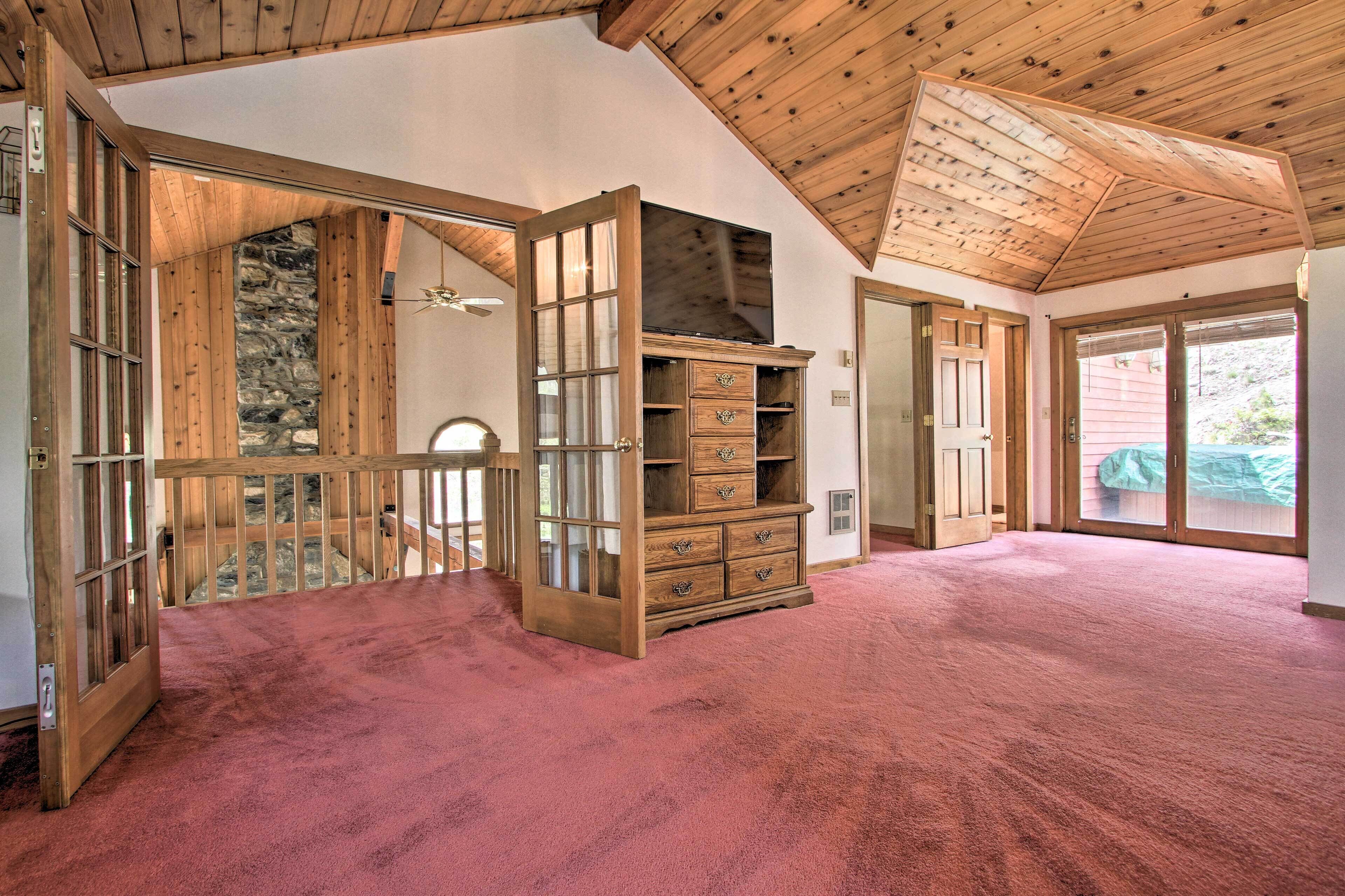 A spacious room with a high ceiling and wooden beams, featuring a large open door leading to a balcony, a built-in wooden cabinet, and a red carpeted floor.