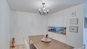 Ein Essbereich mit einem Holztisch, weißen Stühlen, einer Chandelüster und einer Küche im Hintergrund.