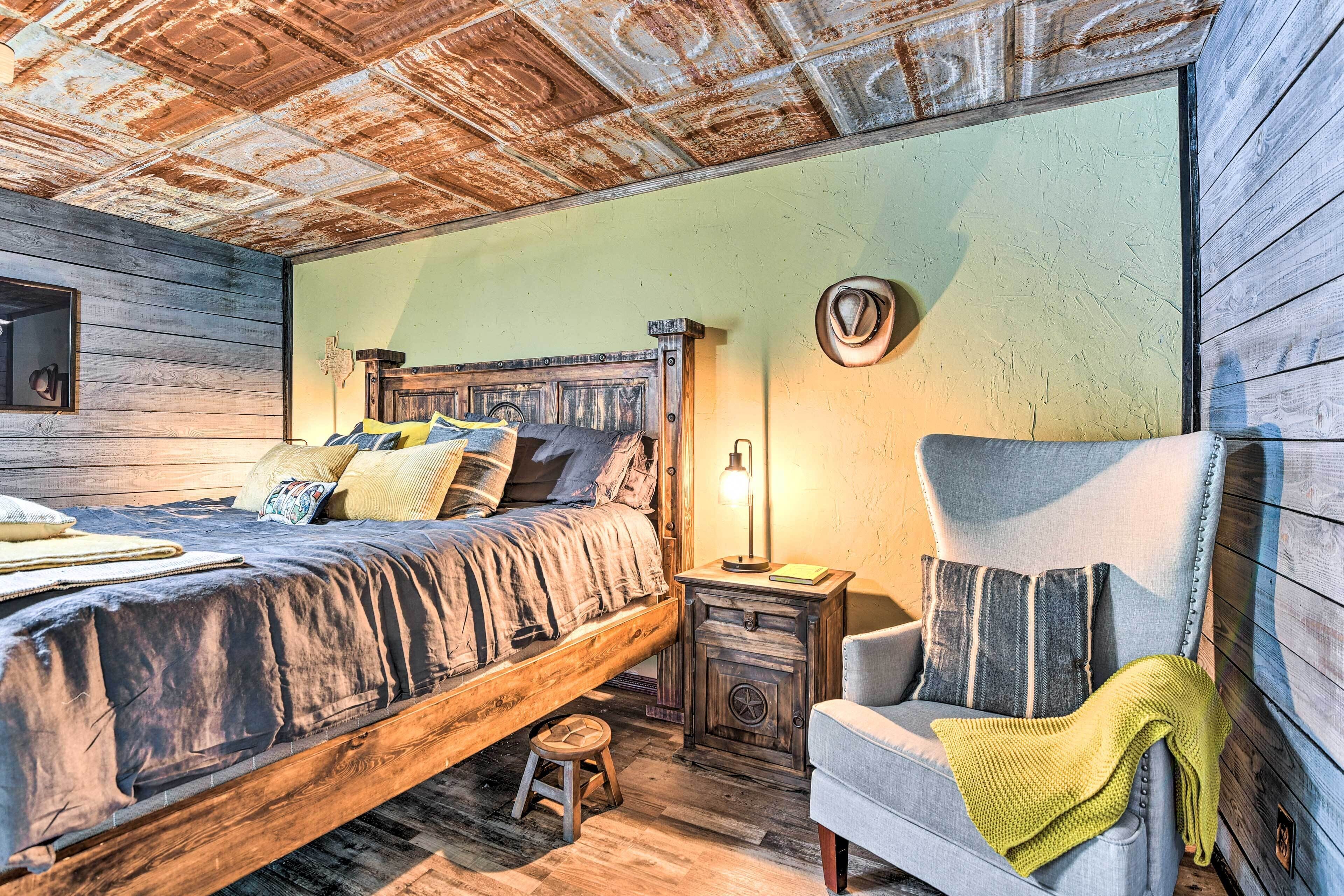 A rustic bedroom with a wooden bed, a small stool, a chair, and a nightstand with a lamp.
