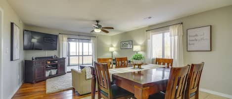 Un comedor con una mesa de madera, sillas y un ventilador de techo. Hay un televisor en la pared y un cartel enmarcado que dice "friends and family gather here".