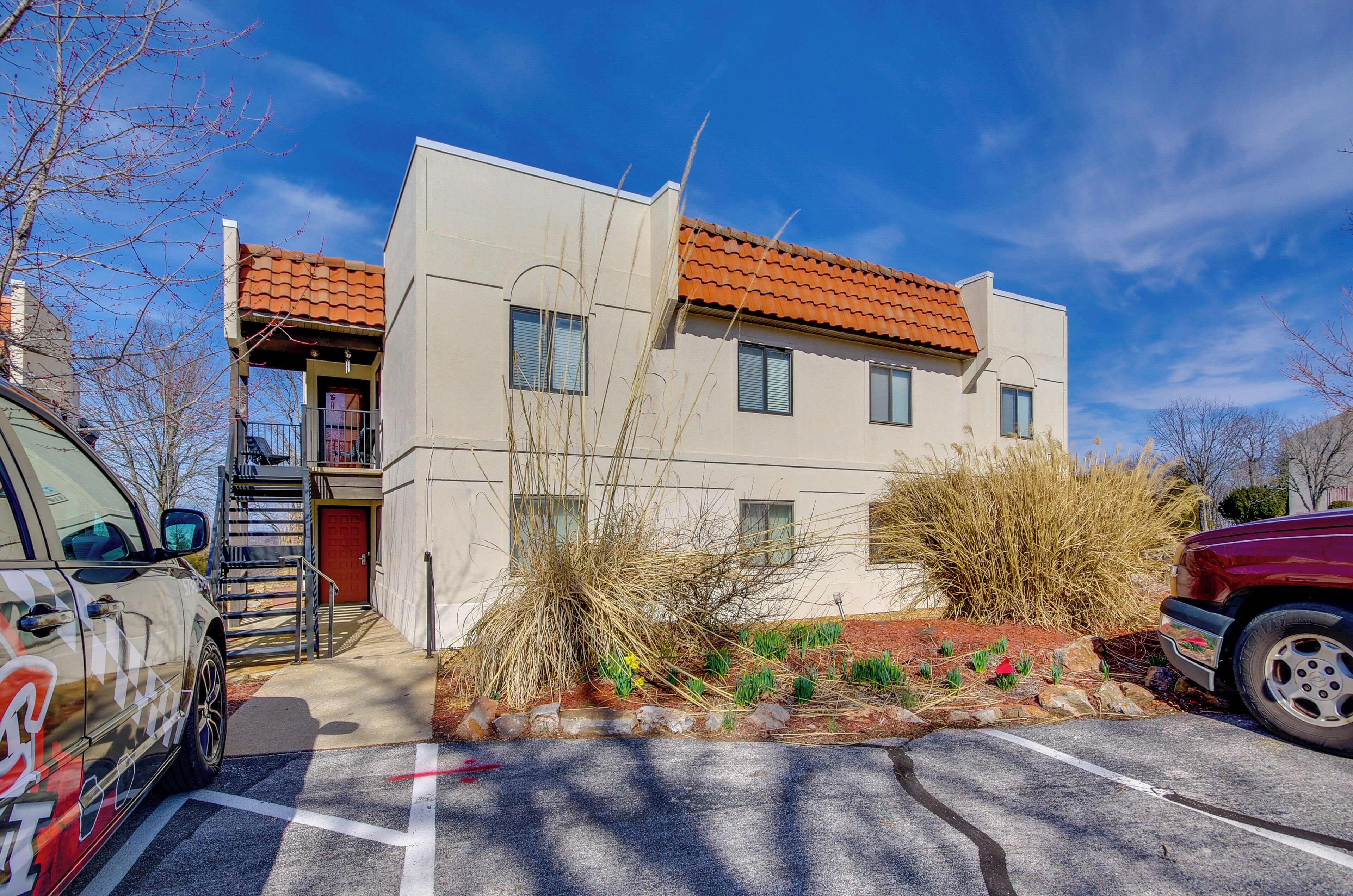 A modern two-story building with a red-tiled roof, a small garden area with plants and rocks, and a parking lot with two cars.