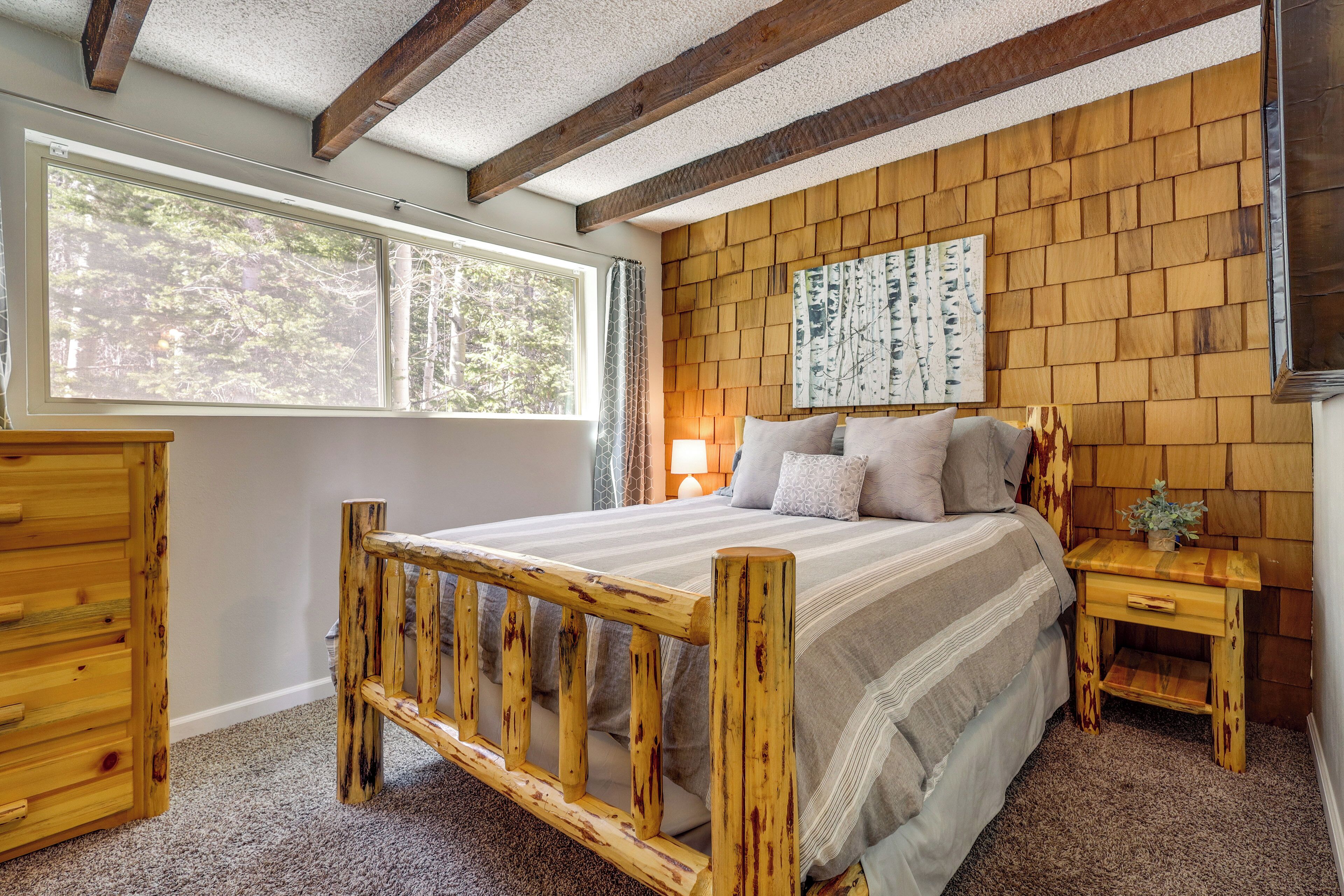 A bedroom with a wooden bed frame, a nightstand, a dresser, and a large window with a view of trees.
