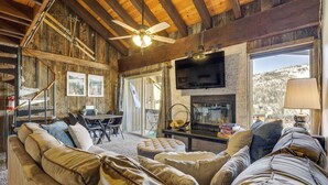 A cozy living room with a fireplace, wooden beams, a ceiling fan, and a large window with a view of the mountains. - Townhome w/ Mtn View - Walk to Brian Head Lifts! (Brian Head)