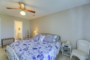 Un dormitorio con cama, una silla de mimbre blanca, una mesita de noche, un ventilador de techo y una puerta que da a otra habitación.