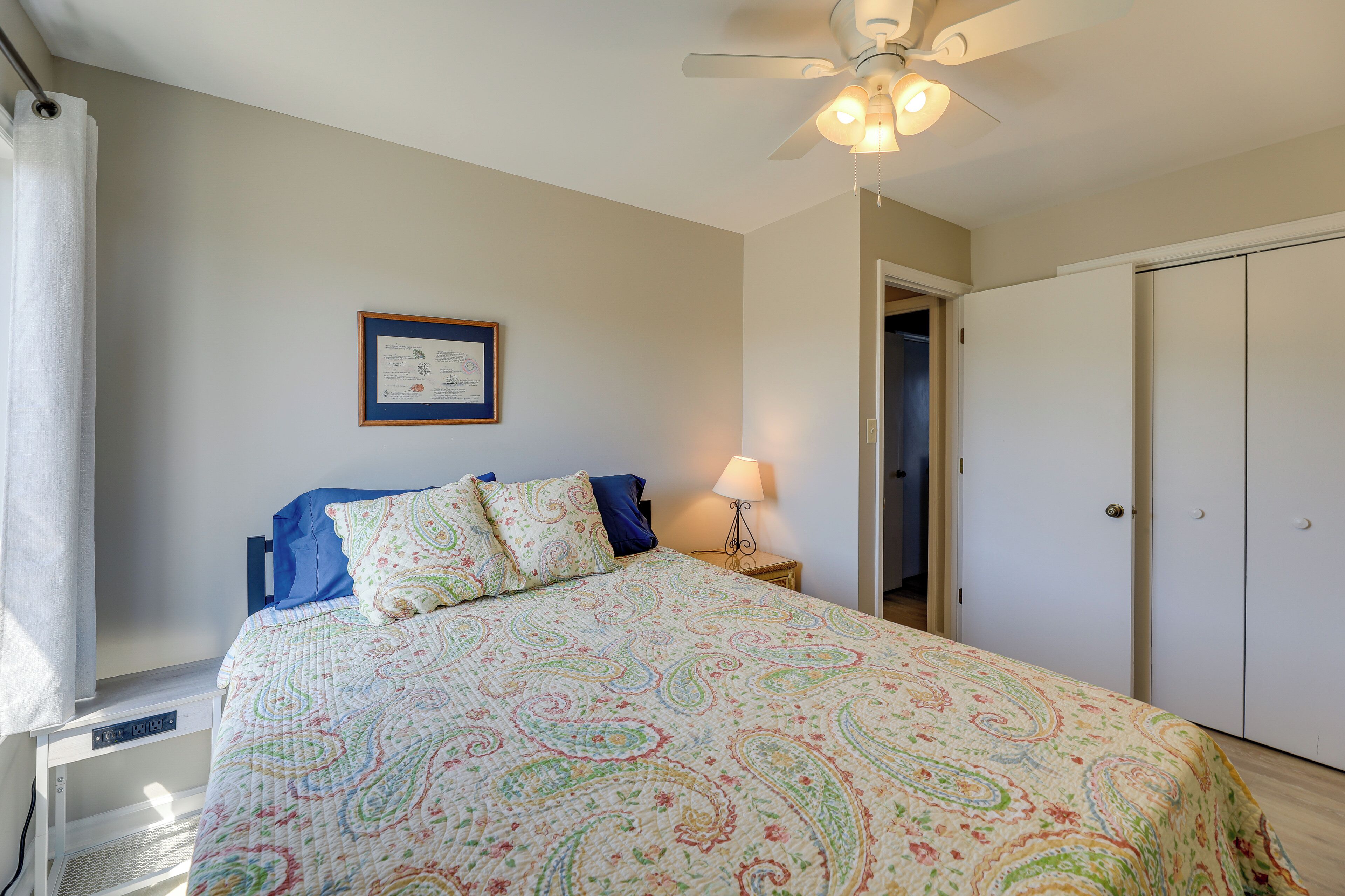 A bedroom with a bed, a ceiling fan, a nightstand with a lamp, a framed certificate on the wall, and a wardrobe.
