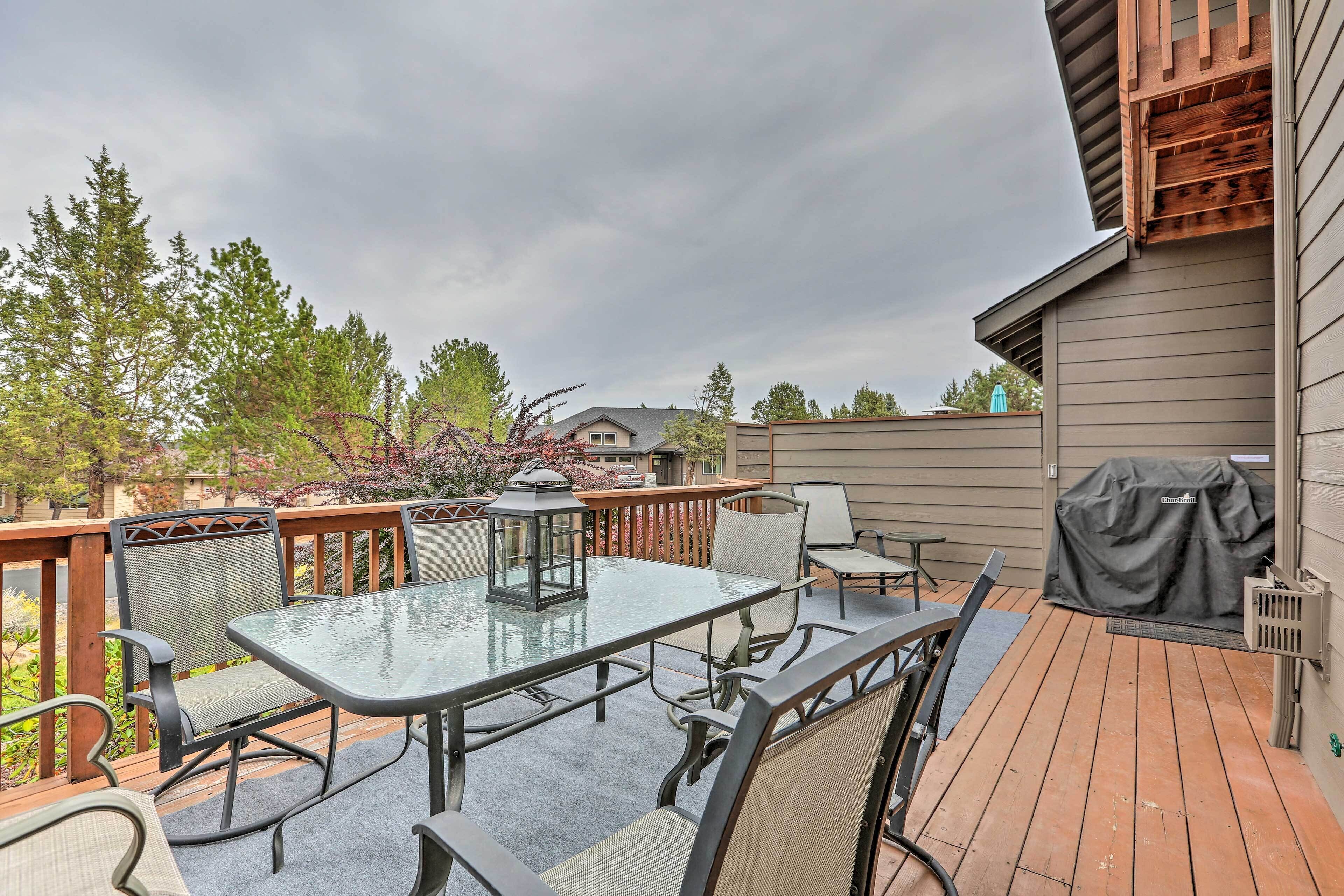 An outdoor patio with a glass-top table and chairs, surrounded by wooden deck railings and a covered grill area.