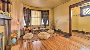 A room with a wooden floor, a ceiling fan, a fireplace, a table with decorative items, and a large window with curtains. - Charming Detroit Abode w/ Meditation Space! (Detroit)