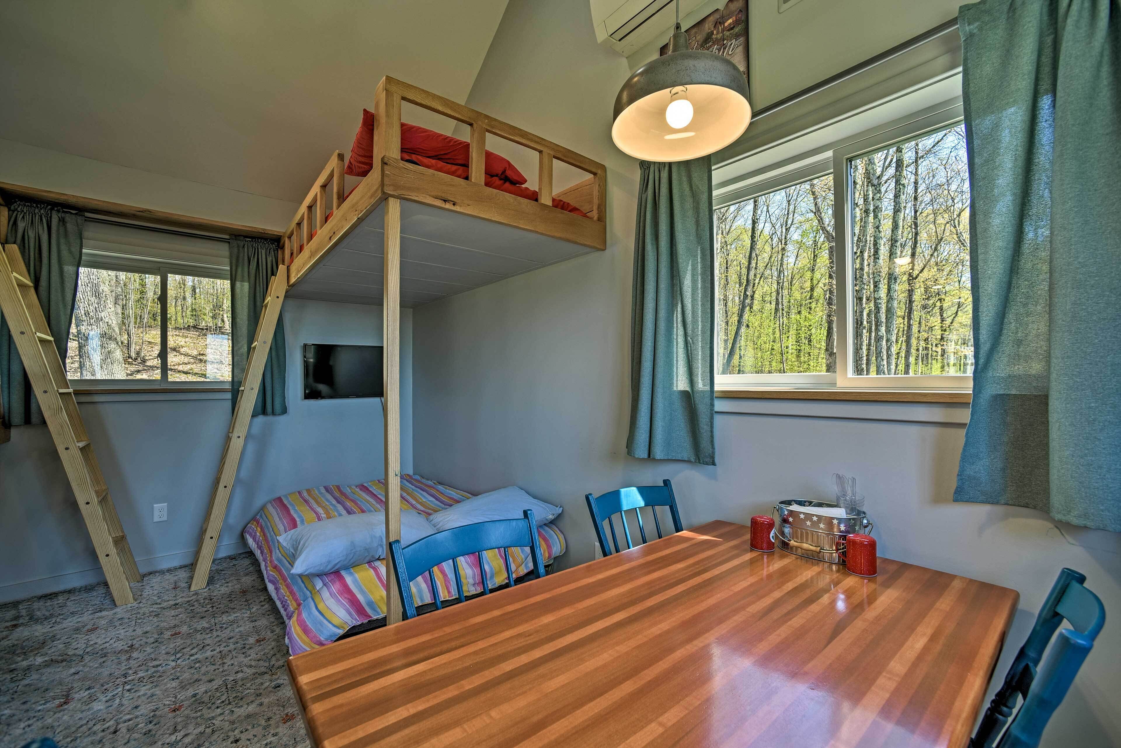 A room with a bunk bed, a dining table, chairs, a lamp, and a view of trees through the windows.