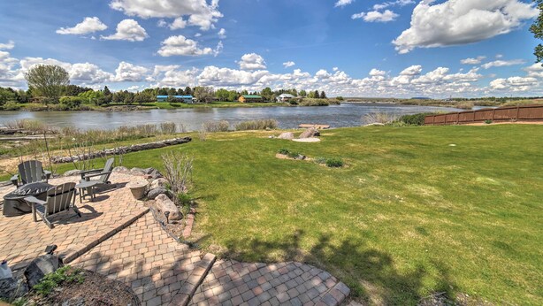 A grassy area with a brick patio, outdoor seating, and a view of a lake and buildings in the distance. - Idyllic Idaho Falls Retreat w/ River Views! (Rigby)