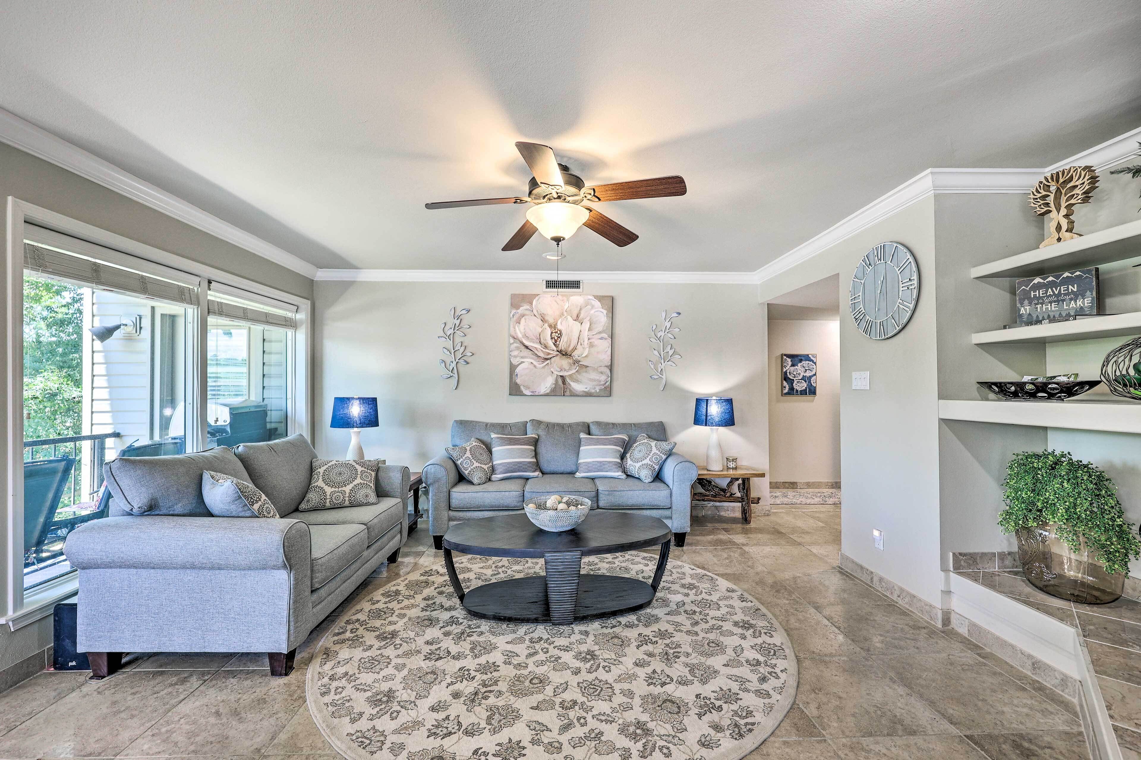 A living room with a sofa, coffee table, and a ceiling fan. There are windows, a painting, and a clock on the wall.