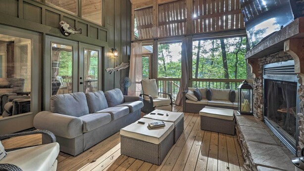 A cozy cabin interior with a fireplace, comfortable seating, and large windows overlooking a forest. - Rustic-chic Riverfront Home w/ Dock, Deck & Canoes (Tallassee)