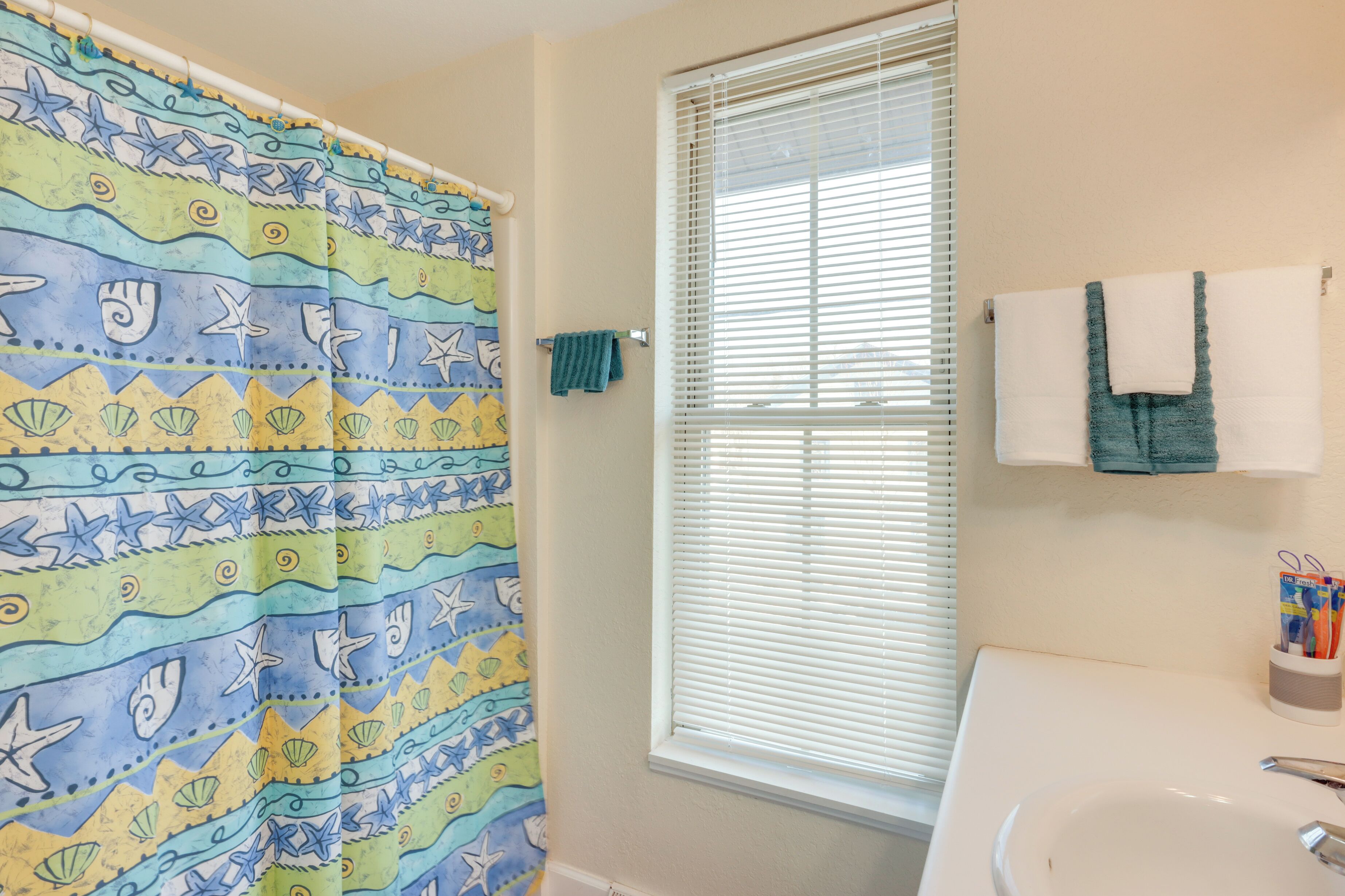 A bathroom with a shower curtain featuring a sea-themed pattern, a window with blinds, a towel rack with a towel, and a sink with a cup of pens.