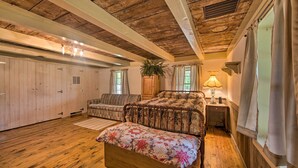 A bedroom with a wooden ceiling, a bed with a floral bedspread, a nightstand with a lamp, a window with curtains, and a potted plant. - Remote 1901 Studio Cabin w/ Loft - Pets Allowed! (Allenwood)