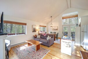 A spacious living room with a sofa, coffee table, and a dining area with chairs and a table. There is a kitchen area with a refrigerator and a window overlooking the outdoors.