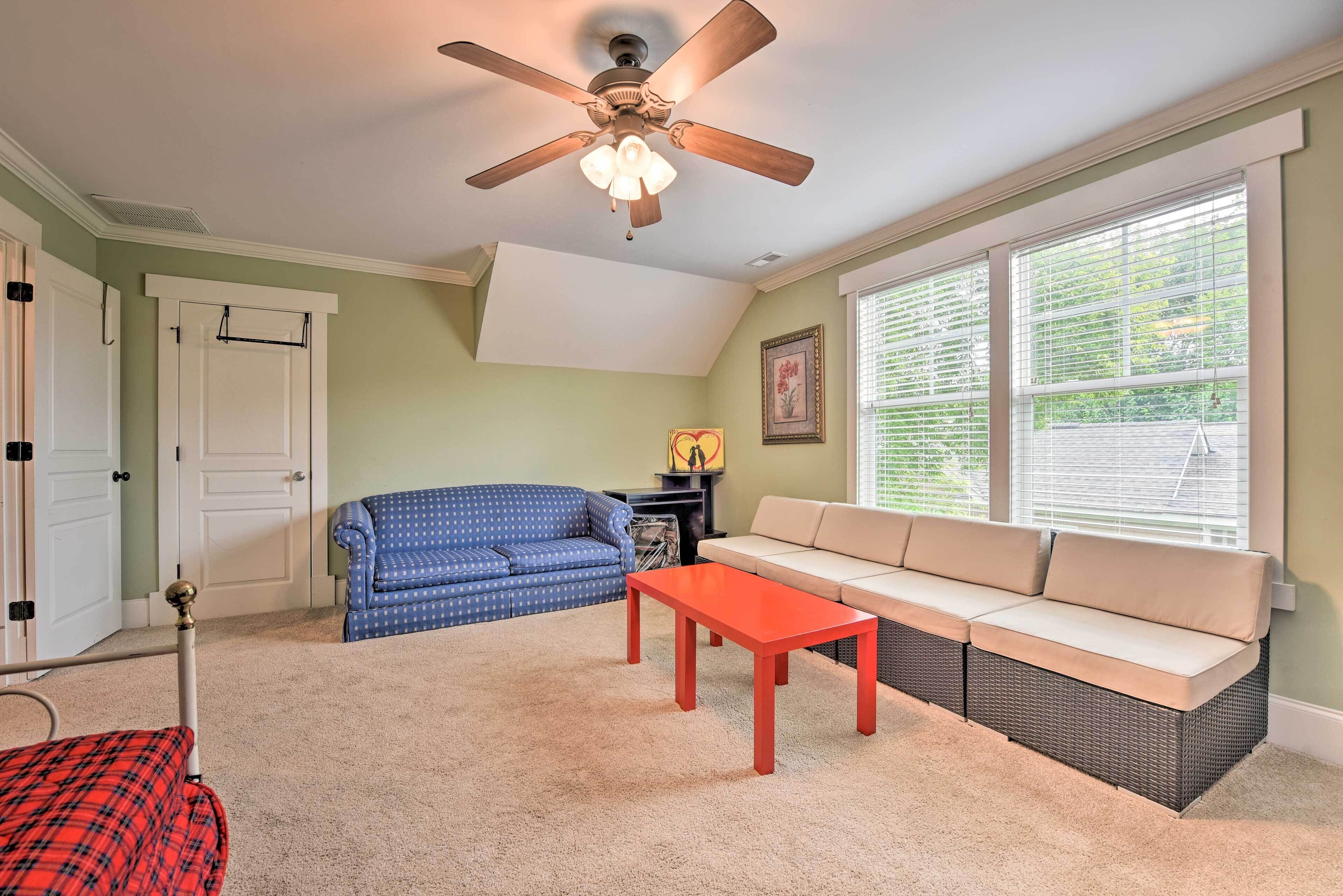 A living room with a ceiling fan, a blue sofa, a beige sofa, a red coffee table, and a large window with blinds.