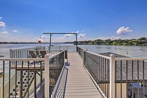 A wooden pier with metal railings, a covered seating area, and a view of a lake with houses in the background. - Lavish Lake House, Near Vineyards + Marinas! (Granbury)