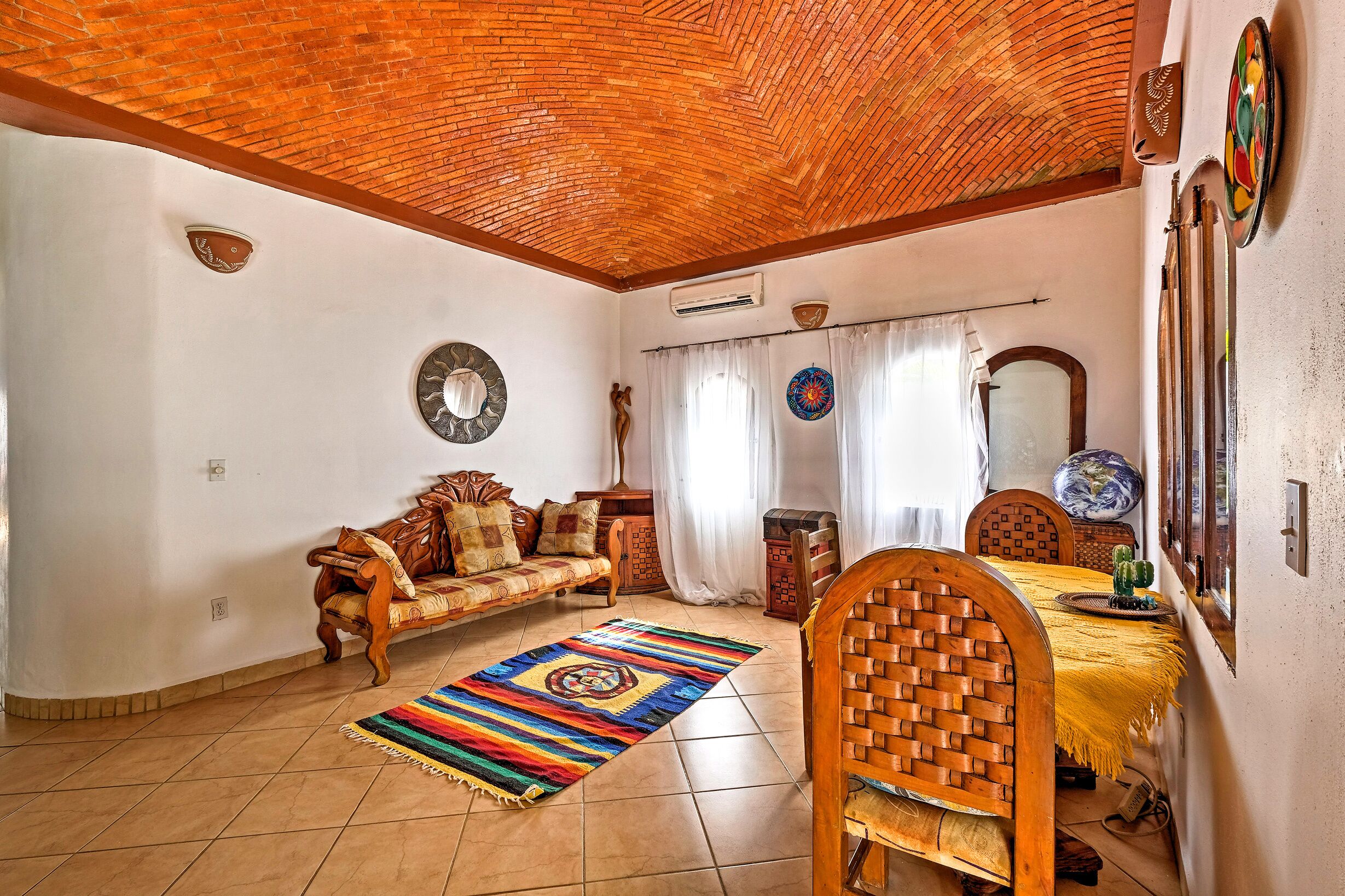 A room with a brick ceiling, a wooden sofa, a dining table with a yellow tablecloth, and a colorful rug.