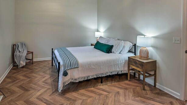 A bedroom with a bed, two bedside lamps, a nightstand, a chair with a blanket, and a herringbone-patterned wooden floor. - Updated Hendersonville Home ~ 3 Mi to Lake! (Hendersonville)