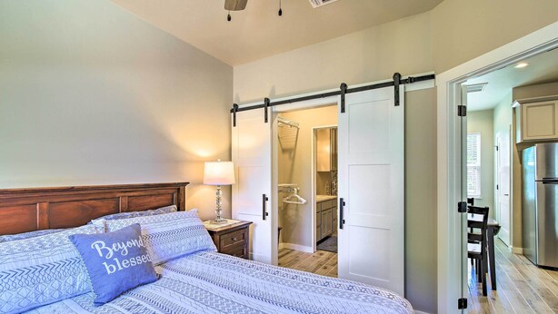 Un dormitorio con cama, una mesita de noche con lámpara, una puerta corredera que da al baño y una cocina visible a través de una puerta abierta.
