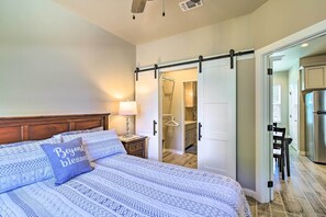 Ein Schlafzimmer mit Bett, Nachttisch mit Lampe, einer Schiebetür zu einem Badezimmer und einer durchgehenden Tür sichtbaren Küche.