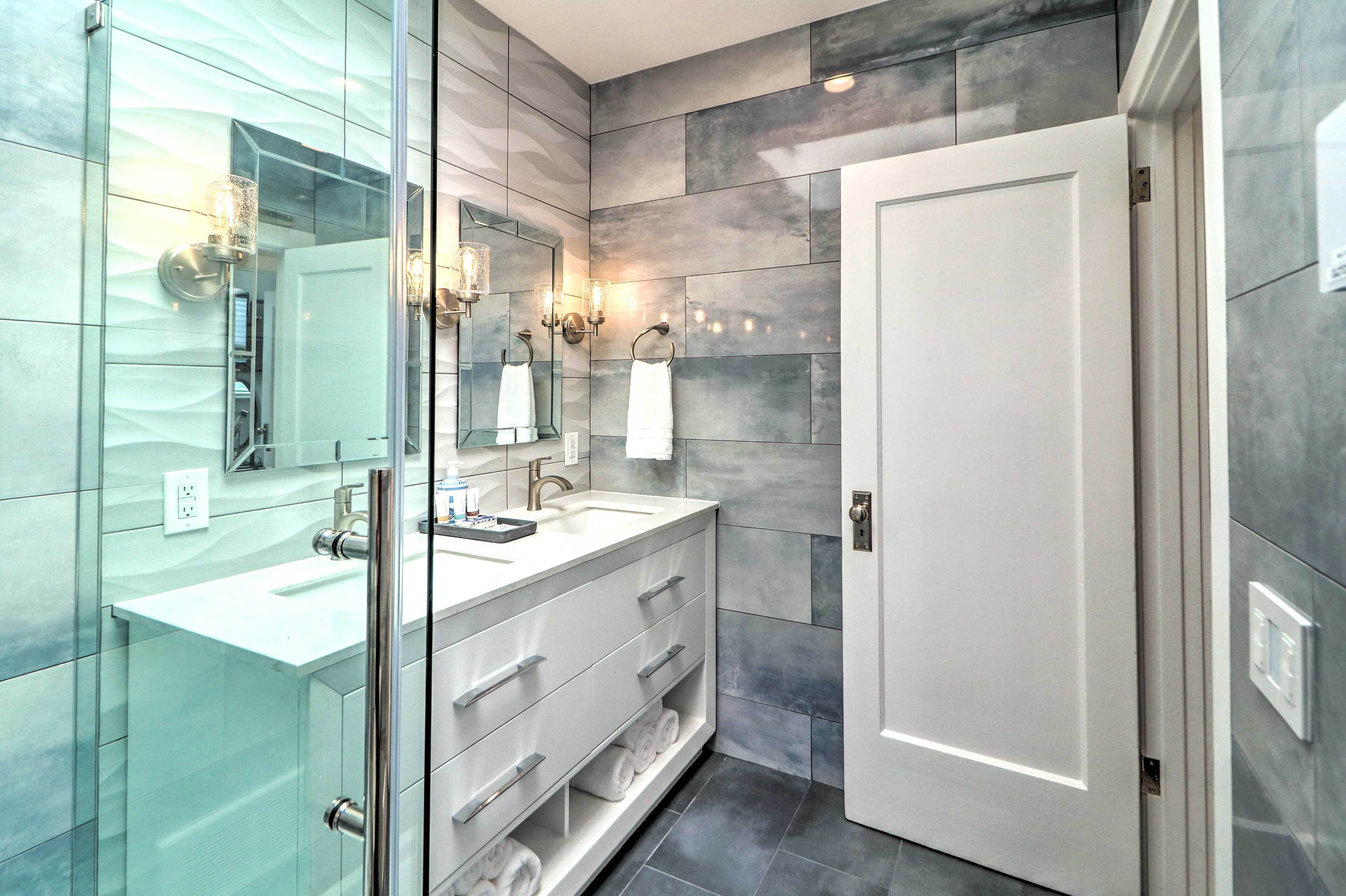 Une salle de bain moderne avec une douche à parois de verre, un meuble-lavabo blanc avec un miroir et une porte dotée d’un interrupteur.