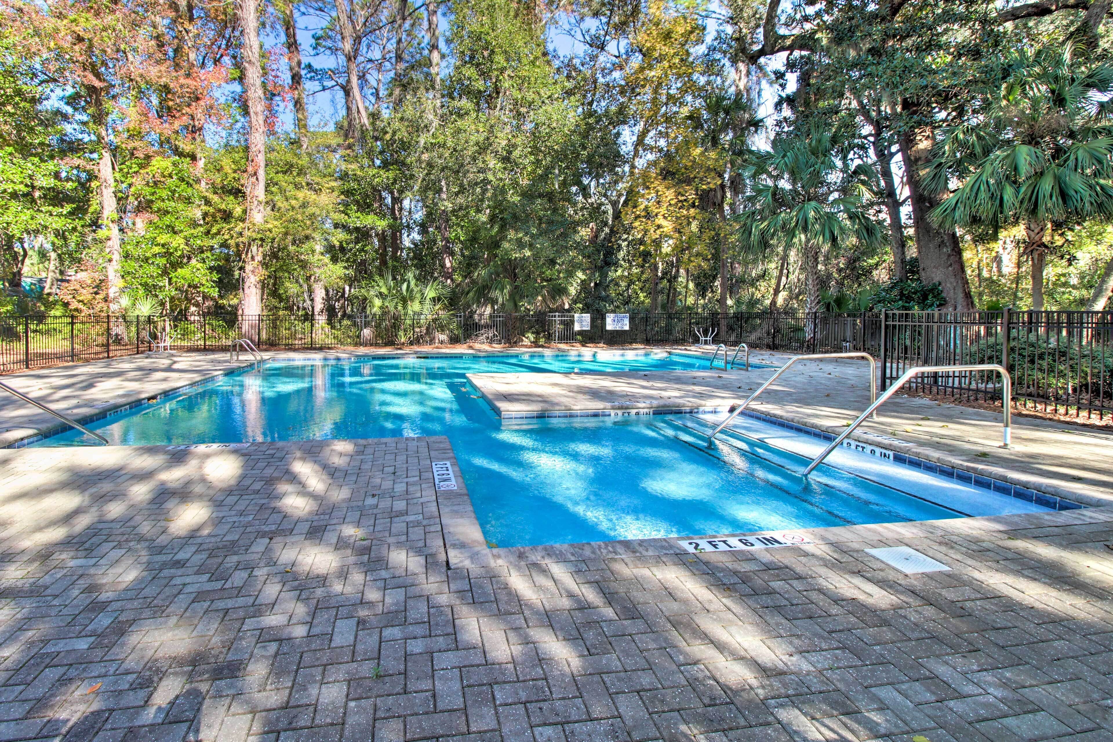 Piscina rectangular con solárium de ladrillo, rodeada de árboles y una cerca de metal.