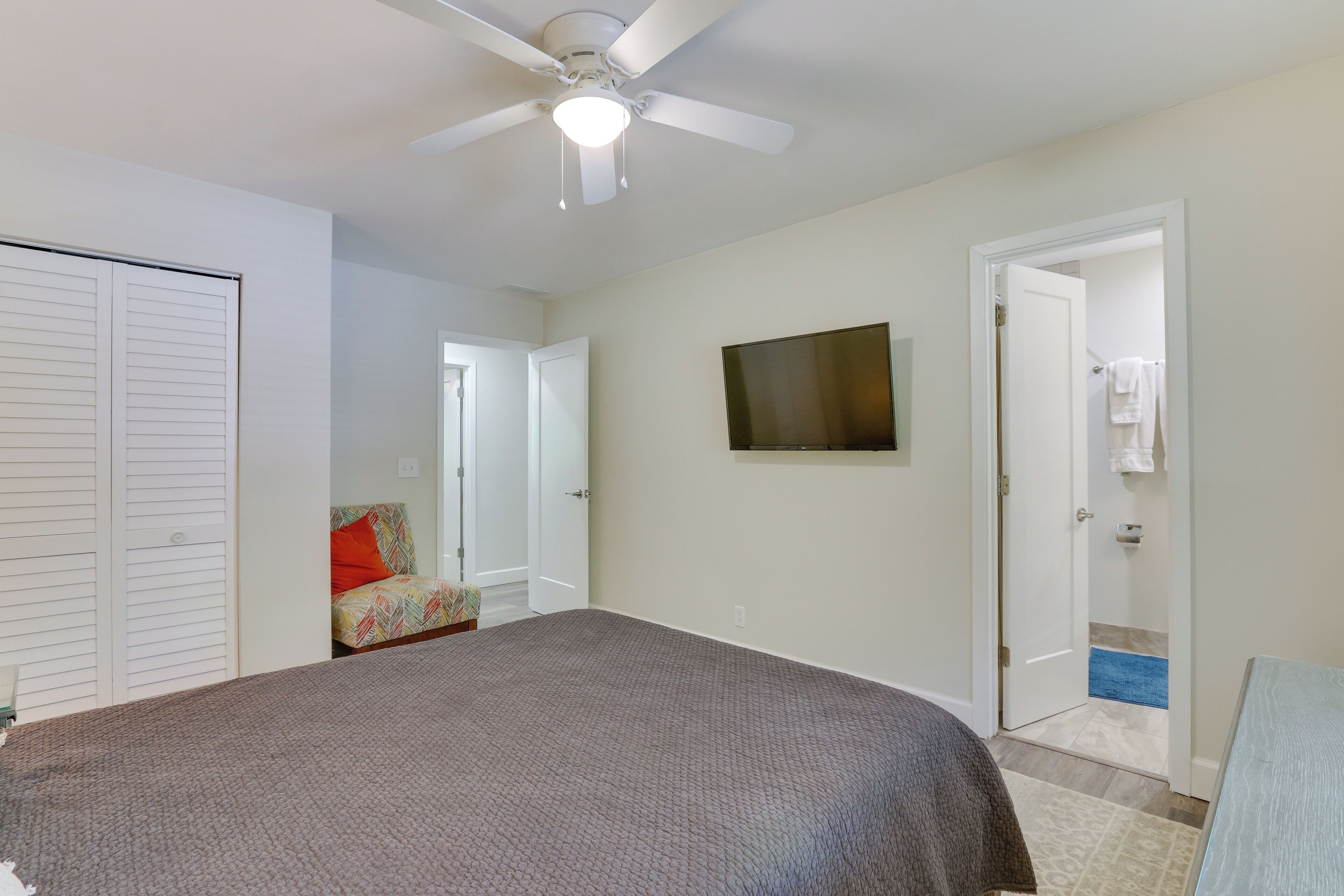 Une chambre à coucher comprenant un lit, un ventilateur de plafond, un téléviseur fixé au mur et une porte donnant sur une salle de bain.
