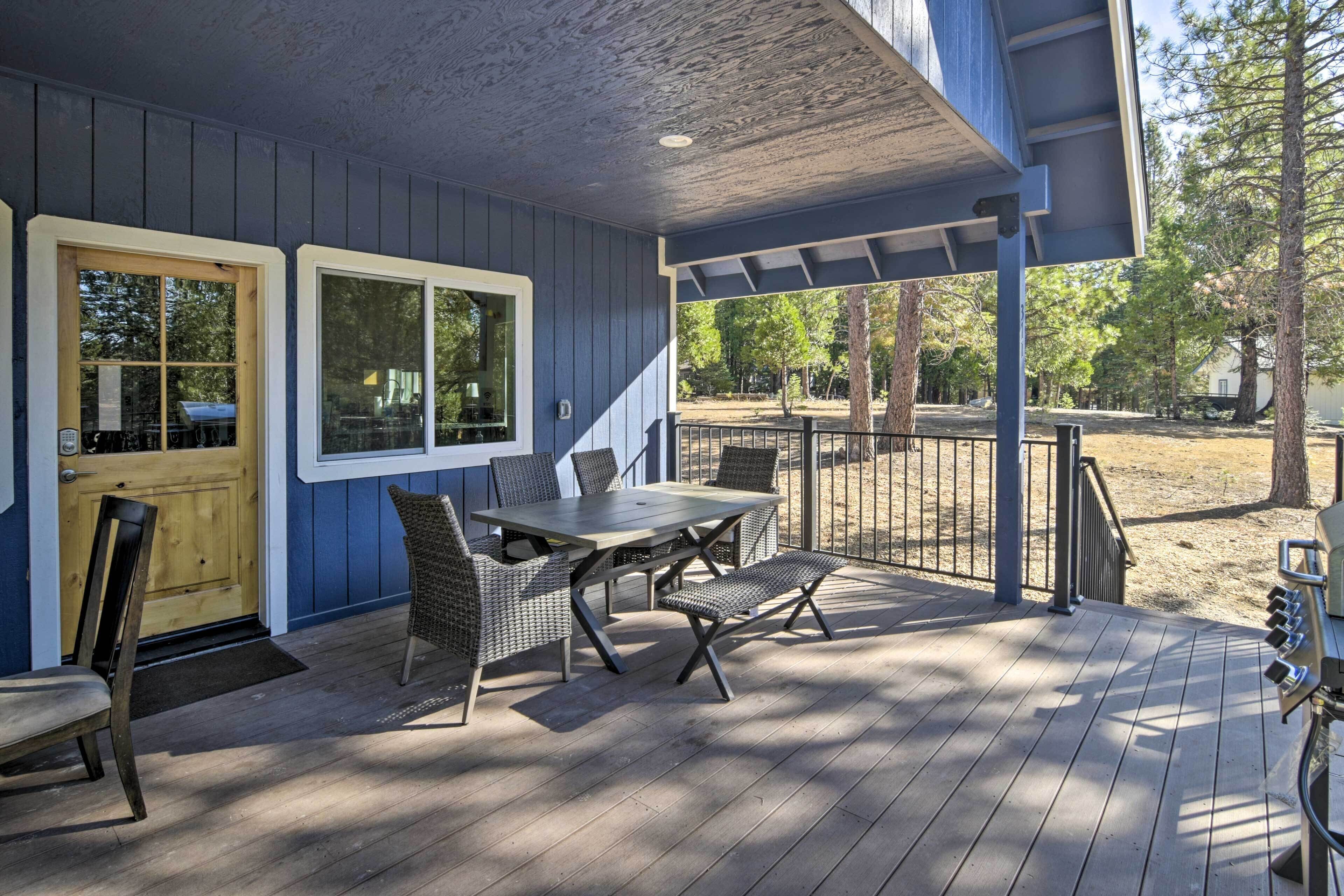 A covered outdoor patio with a table and chairs, a wooden door, and a view of trees and a building in the background.