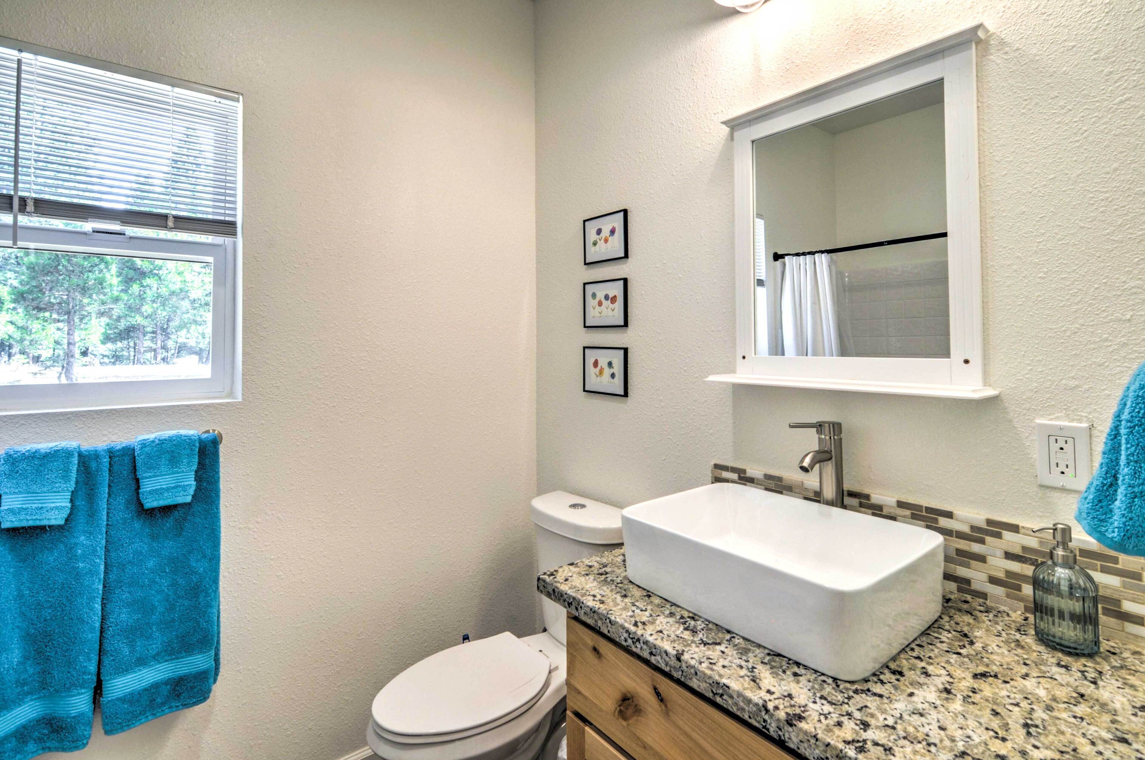 A bathroom with a white sink, a toilet, and a window with blinds. There are three framed pictures on the wall and a blue towel hanging on the wall.