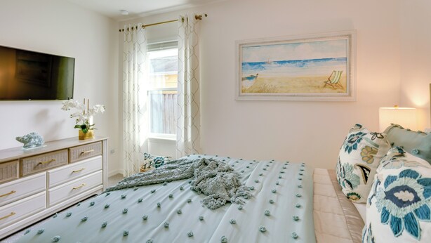 Une chambre à coucher avec un lit, une commode, un téléviseur, un tableau et une fenêtre avec des rideaux.