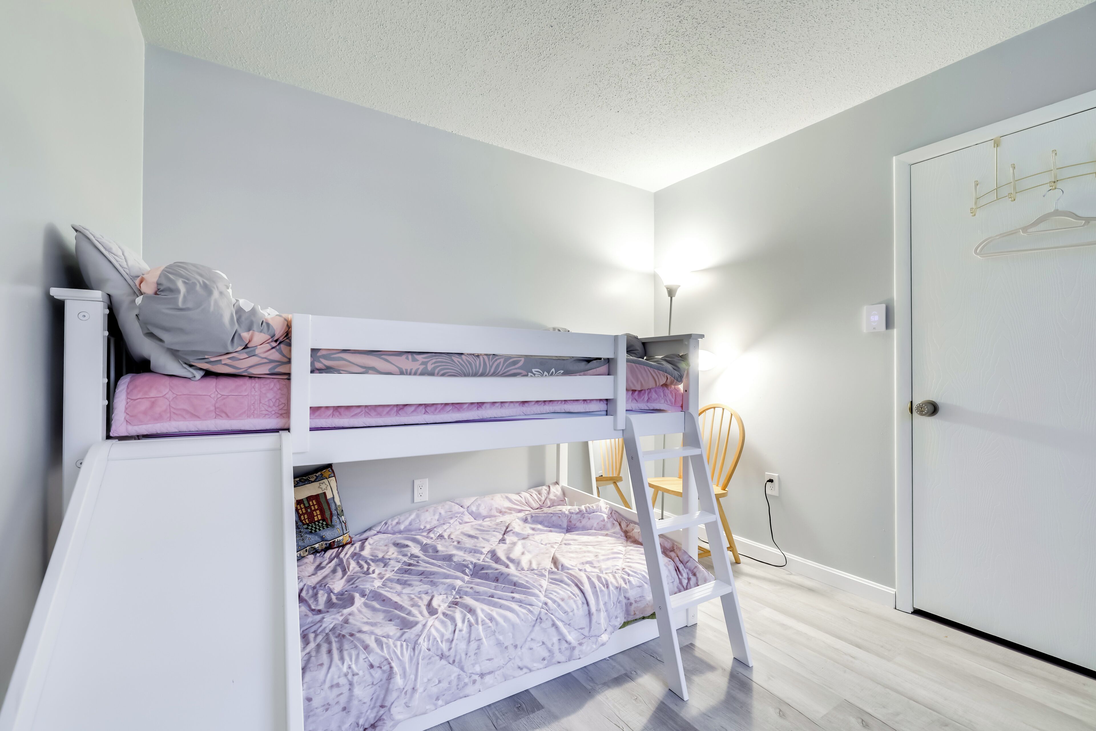 Un lit superposé avec une échelle, un bureau et une chaise, dans une pièce aux murs de couleur claire et dotée d’une porte.