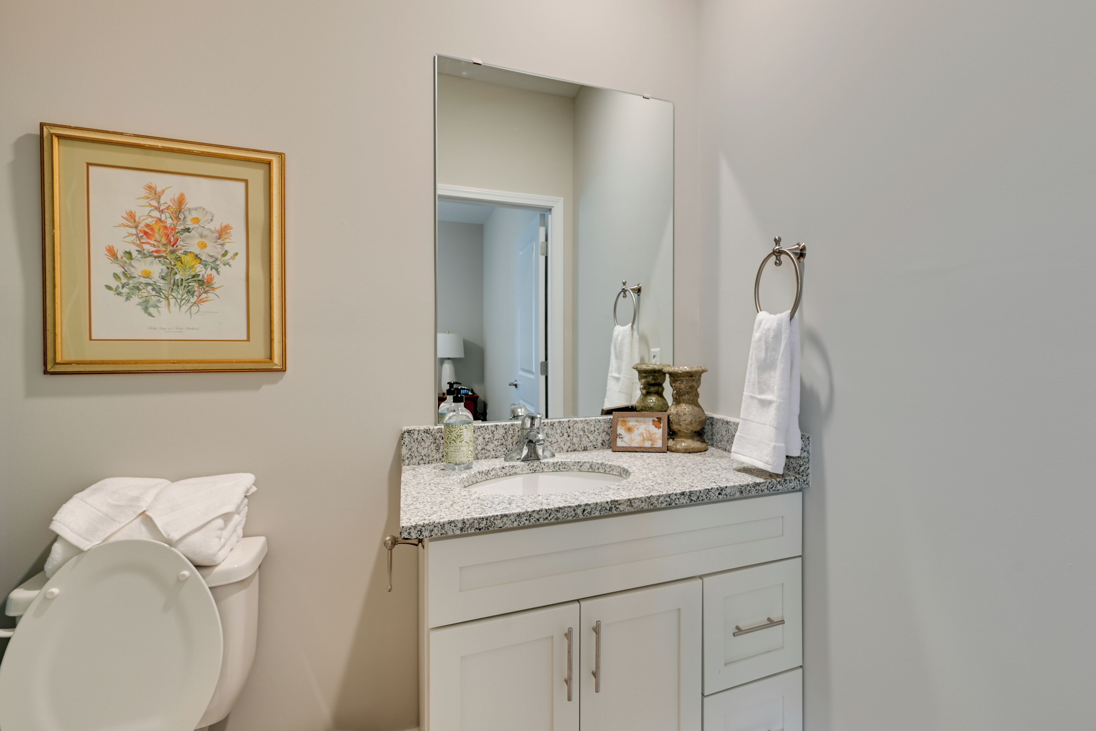 A bathroom with a white toilet, a sink with a marble countertop, a large mirror, and a framed floral artwork on the wall.