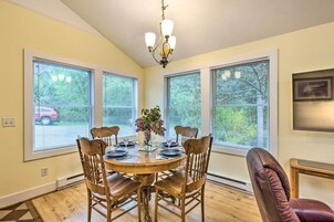 A dining room with a round wooden table set for four, surrounded by wooden chairs. There is a chandelier above the table, and the room has large windows with blinds.